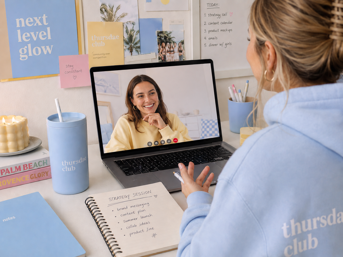 A woman in a light blue hoodie with 'thursday club' printed on the back sits at a desk, video calling another woman on a laptop. The desk is decorated with a blue tumbler, a candle, and notebooks under a whiteboard with weekly goals.